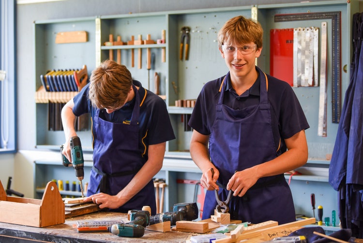 Two students in a woodwork shop one with a drill and the other looking at the camera.